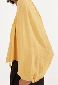 Żółta bluzka o fakturowanej powierzchni z luźnymi, puffed rękawami; posiada plisowany wzór i elastyczny dół. Noszona na czarne spodnie.