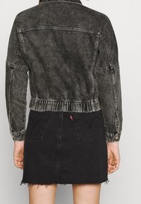 Osoba oblečená ve vybledlé černé džínové bundě a černé džínové sukni s zadními kapsami a roztrhaným lemem, stojící s uvolněnýma rukama.