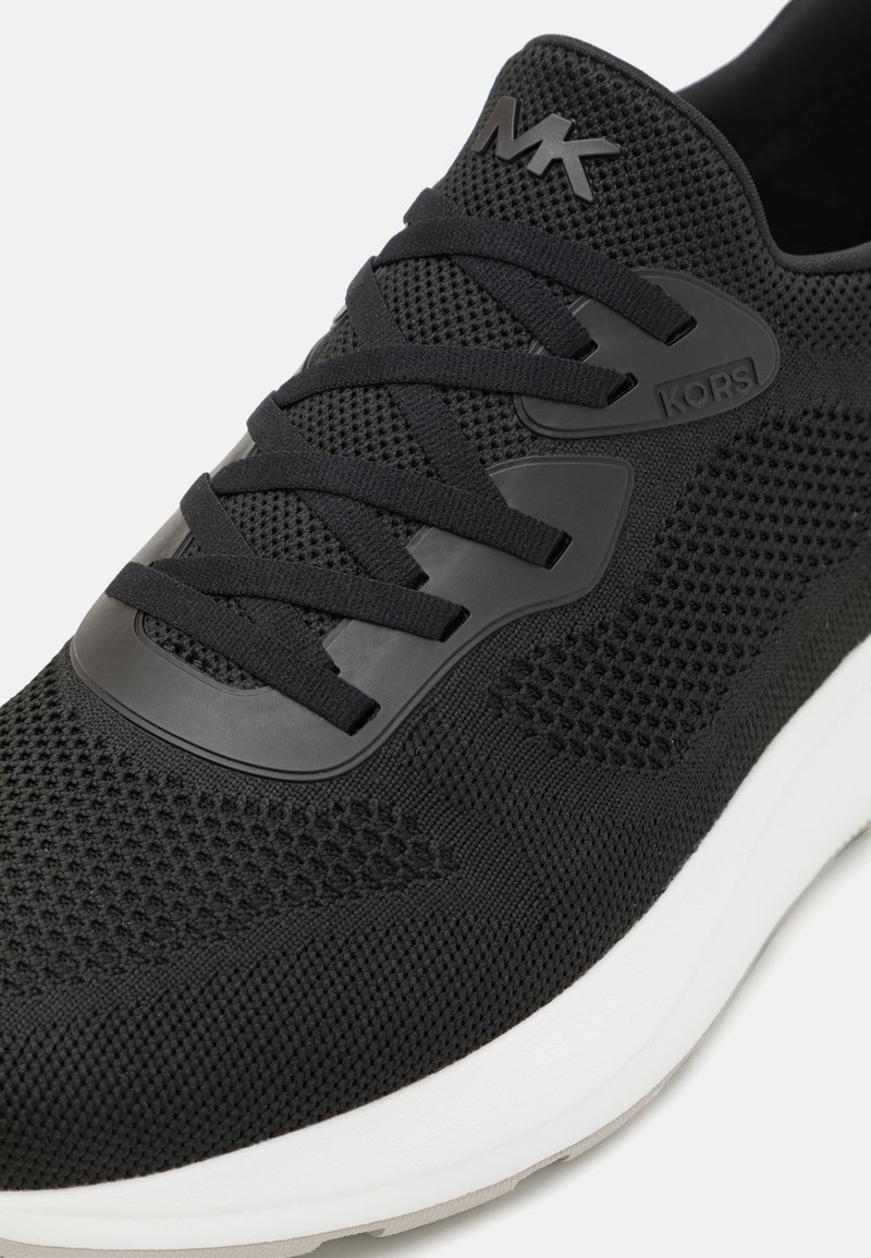 Schwarzer Sportschuh mit Mesh-Obermaterial, geprägtem Logo, flachen Schnürsenkeln und weißer Außensohle. Texturierte Details verbessern den Halt und die Atmungsaktivität.