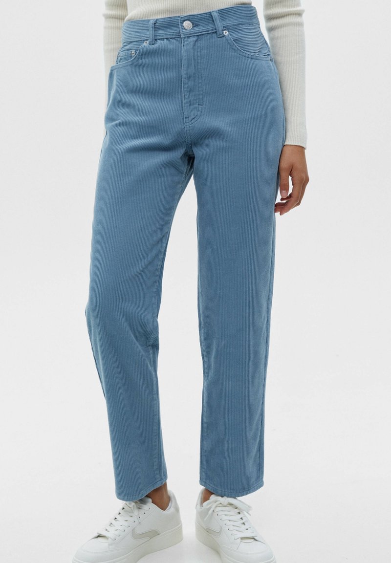Pantalon en velours côtelé bleu clair avec taille haute, coupe droite, coutures visibles et poches avant, associé à des baskets blanches.