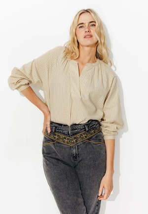 Chemise beige à manches longues et en côte avec un décolleté en V plongeant, associée à un jean en denim foncé taille haute avec des broderies sur la ceinture.