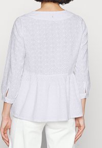 Weiße Lochspitzenbluse mit 3/4-Ärmeln und Peplum-Saum, von hinten getragen, kombiniert mit weißen Hosen.