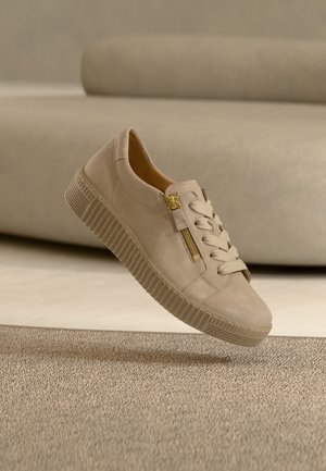 Beiger Freizeitschuh mit dicker Sohle, Schnürung vorne und seitlichem Reißverschluss, auf einem beigen Teppich vor einem beigen Sofa platziert.