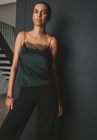 Dunkelgrünes Satin-Cami mit transparenter Blumen-Spitze am Ausschnitt, kombiniert mit hoch taillierten schwarzen Hosen. Einfache Träger und lockere Passform.