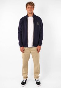 Veste zippée marine avec col haut, portée sur un t-shirt blanc, associée à un pantalon kaki et des baskets noires. Présente un subtil détail de broderie.