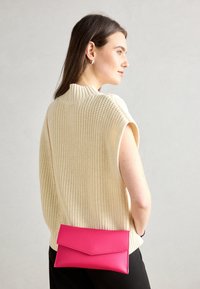 Žena s dlouhými hnědými vlasy, oblečená v krémovém bezrukávovém pleteném topu a černých kalhotách, držící jasně růžovou crossbody kabelku.