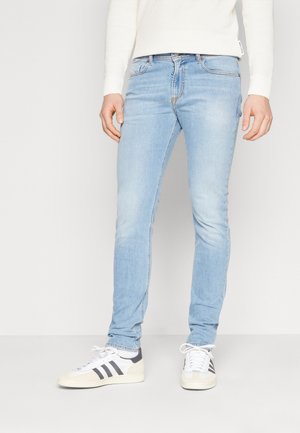 Homme portant un jean slim bleu clair, un haut à manches longues blanc et des baskets blanches avec des rayures noires, debout sur un fond blanc.