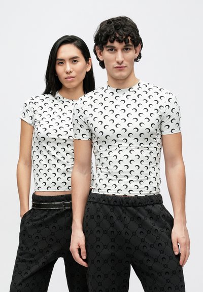 T-shirts brancas justas com padrão repetido de luas crescentes negras, combinadas com calças pretas que apresentam um design subtil e cinto com detalhes de tachas.