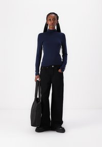Haut à manches longues bleu marine avec un col haut et des accents de boutons aux poignets, associé à un pantalon noir à jambes larges et un sac fourre-tout noir.