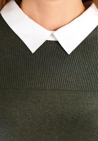 Olivgrön tröja med ruggad textur och vit platt krage. Närbild på krag- och halslinjedetaljer.