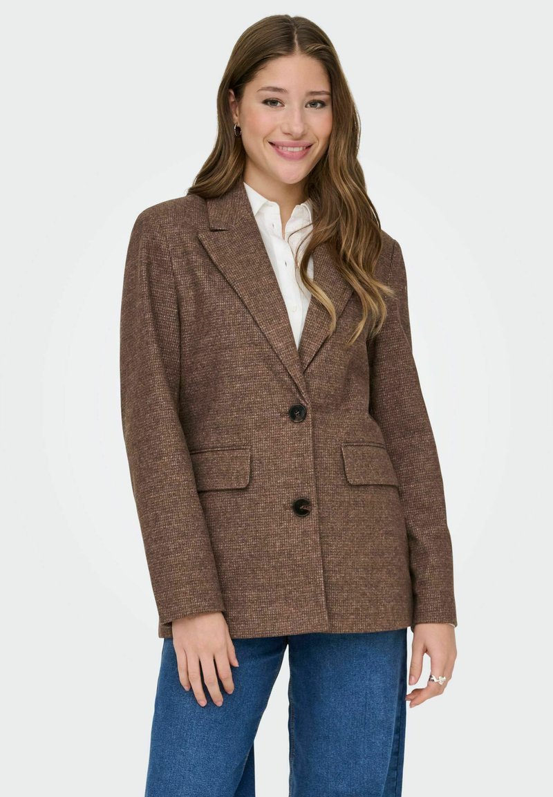 Bruine, gestructureerde blazer met geknipte revers, twee voorkruiszakken en een enkele knoopsluiting, gecombineerd met een wit shirt en blauwe jeans.