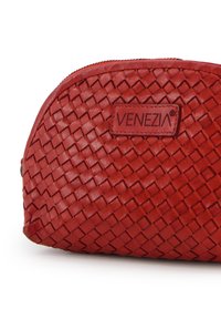 Červená tkaná kožená taška zaobleného tvaru. Má boční zip a obdélníkový štítek s vyraženým nápisem "VENEZIA." Hladký povrch.