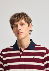Bordeaux- och vitrandig polo shirt med en marinblå krage, med en liten blå logotyp på bröstet och knapplista vid halsen.