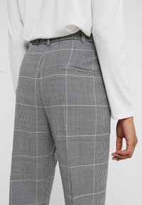 Grijze geruite broek van middelzwaar materiaal, met zwart-witte ruiten en subtiele perzikkleurige accenten; zakken zichtbaar aan de achterkant.