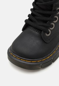 Bota de cuero negra hasta el tobillo con punta redonda, superficie texturizada, cordones negros, ojales metálicos y una costura amarilla distintiva a lo largo de la suela.
