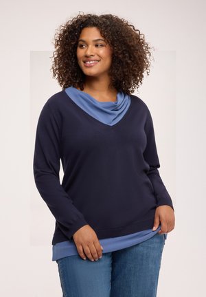 Kvinde med krøllet hår, smilende, iført navyblå V-hals sweater over lyseblå top og blå jeans, stående mod en lys baggrund.