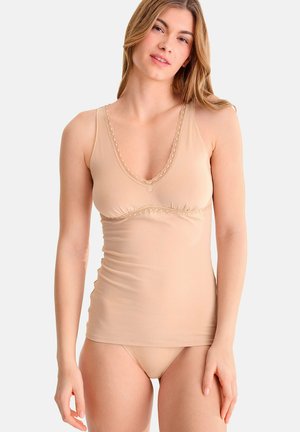 Femme portant un haut beige sans manches avec bordure en dentelle et sous-vêtements beige assortis, debout devant un fond clair uni.
