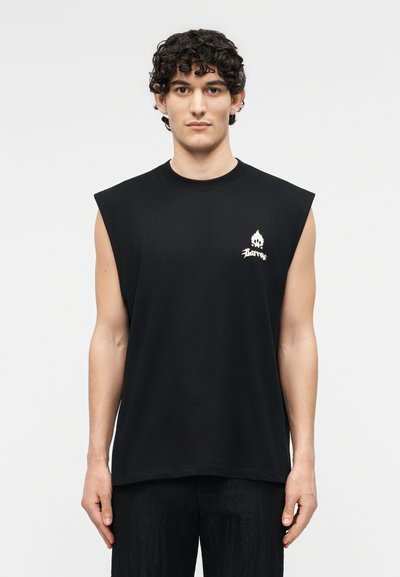 Νέος άνδρας με σγουρά μαλλιά, φοράει αμάνικο μαύρο πουκάμισο με ένα μικρό λευκό λογότυπο φλόγας και κείμενο στο στήθος, πάνω σε απλό φόντο.