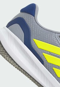 Zapatilla atlética gris con rayas amarillas, acentos en azul marino y parte superior de malla texturizada. Media suela blanca con una suela exterior gris estriada.