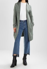 Kvinna som bär en lång grön hoodiekofta, vit T-shirt, korta blå jeans och svarta klackade ankelskor, stående mot en neutral bakgrund.