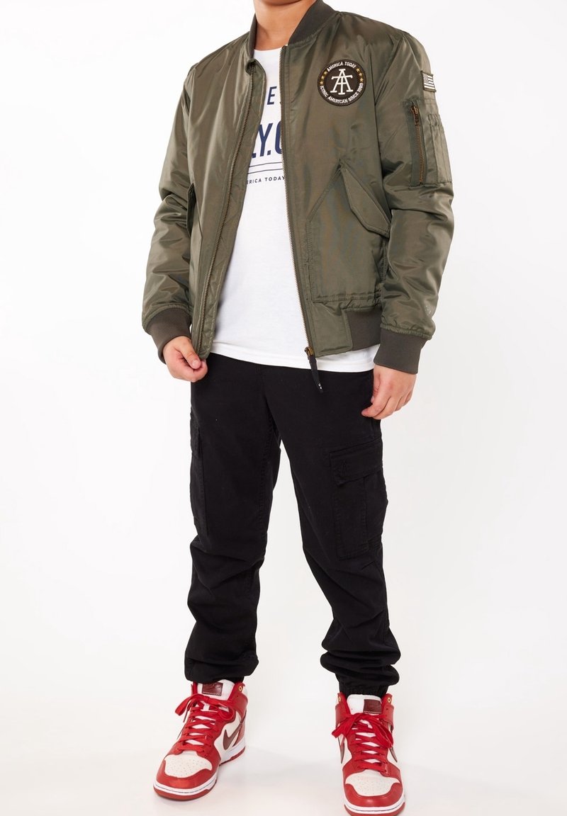 Veste bomber vert olive avec un patch rond, poches zippées, poignets et ourlet côtelés. Associée à un pantalon cargo noir et des baskets rouges.