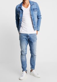 Man in een lichtblauwe spijkerjas, een witte V-hals T-shirt, strakke lichtblauwe jeans en witte sneakers voor een effen achtergrond.