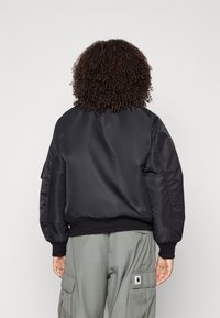 Zwarte bomberjack met een gladde textuur, ribgebreide manchetten en zoom, en details van zijzakken. Gecombineerd met lichtgroene cargobroeken.