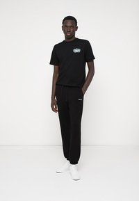 Černé tričko s grafickým logem a černé tepláky. Oba kousky mají volný střih. Celý outfit doplňují bílé tenisky.