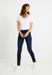 Camiseta blanca de cuello en V combinada con vaqueros pitillo azul oscuro y zapatillas blancas. La camiseta tiene mangas cortas y un ajuste informal.