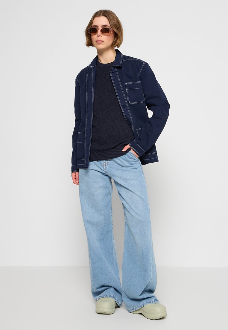 Granatowa, lekka kurtka z kontrastowym przeszyciem, zestawiona z ciemnym swetrem, szerokimi jasnoniebieskimi dżinsami oraz beżowymi butami wsuwanymi.