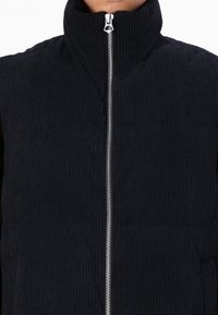 Zwarte corduroy vest met een hoge kraag en zilveren rits. De stof heeft een ribbelstructuur en een strakke, minimalistische uitstraling.