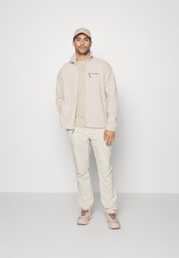 Lys beige fleecejakke med lynlås, båret over en lys sandfarvet skjorte, parret med lys beige bukser og beige træningssko. Kappen matcher outfittet.