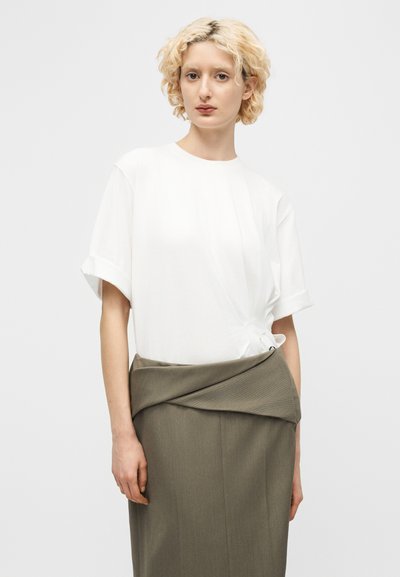 Γυναίκα με κοντά σγουρά ξανθά μαλλιά, που φοράει λευκό τοπ με κοντά μανίκια και γκρι φούστα ψηλόμεση με διπλωμένη μέση.