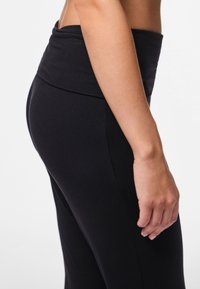 Zijaanzicht van een persoon die hoog getailleerde zwarte leggings draagt, waarbij een deel van de romp, arm en hand zichtbaar is tegen een witte achtergrond.
