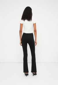 Witte aangesloten t-shirt en hooggetailleerde zwarte flared broek met een kettingaccent op de taille; model staat met de rug naar de kijker.