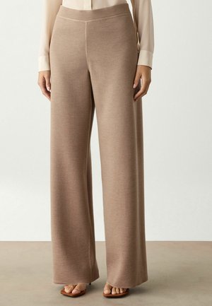 Kvinde iført beige strikkede bukser med høj talje og vide ben, med en lysebeige bluse stoppet ned i bukserne og åbne sandaler.