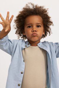 Camicia azzurro chiaro con maniche arrotolate sopra un body beige a coste. Il bambino mostra cinque dita con una mano e fa il broncio.