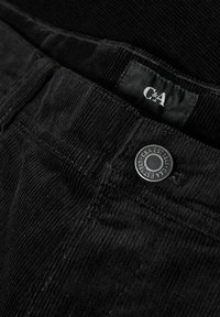 Gros plan sur un pantalon en velours côtelé noir avec un bouton en métal et une étiquette en tissu noir cousue portant le logo "C&A".