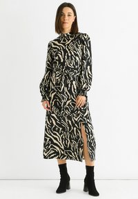 Long black and cream dress with a zebra pattern, high neck, belted waist, long sleeves, and a thigh-high slit. Paired with black ankle boots.