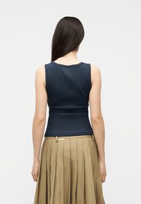 Femme aux longs cheveux foncés portant un haut sans manches bleu marine à côtes et une jupe plissée beige, vue de dos sur un fond uni.