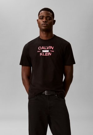Homme portant un t-shirt noir Calvin Klein avec un logo rose, un pantalon noir et une ceinture noire, debout devant un fond gris uni.