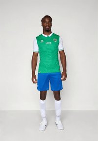 Camisola de futebol verde com mangas brancas, calções azuis e meias brancas com padrão de pontos. A camisola apresenta um logo e detalhes em texto.
