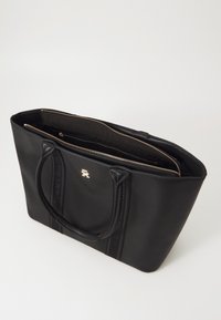 Bolso tote negro hecho de material liso que cuenta con doble asa, compartimentos con cremallera y un logo dorado en la parte frontal. Interior forrado en negro.