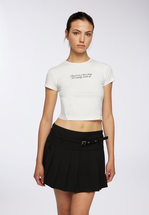 T-shirt bianco corto con maniche corte e design testuale, abbinato a una gonna nera plissettata con cintura e dettagli frontali.