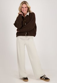 Bruin gebreid vest met grote knopen over een wit shirt; crèmekleurige wijd uitlopende broek; bruine schoenen met witte strepen. Gladde textuur.