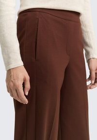 Braune maßgeschneiderte Hose mit glatter Textur, seitlichen Taschen und geradem Bein Design. Der Bund ist flach und minimalistisch.