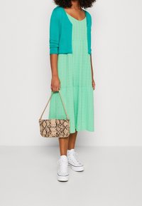 Šaty v mátové barvě, se sklady a texturovaným povrchem, doplněné aquamarínovým cardiganem. Drží vzorovanou tašku s b�éžovým hadím potiskem. Bílé tenisky.