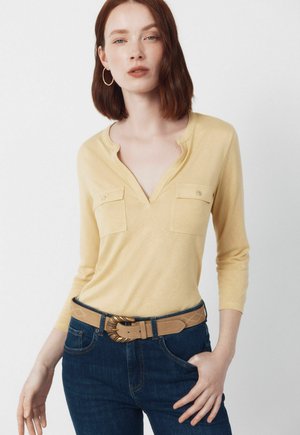 Młoda kobieta z czerwonymi włosami nosi bladoróżowy top z dekoltem w kształcie litery V i kieszeniami na piersi, niebieskie dżinsy oraz beżowy pasek z ozdobną złotą klamrą.