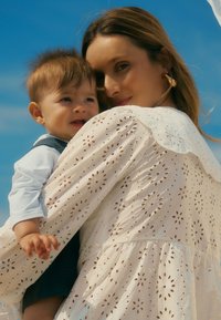 Witte jurk met gaatjespatronen en een gekartelde kraag, vastgehouden door een persoon die een baby in een blauwe outfit draagt. Blauwe lucht op de achtergrond.