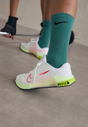Personne portant des baskets Nike blanches et vert fluo et des chaussettes Nike turquoise, tenant une barre, se préparant à soulever des poids à l’intérieur.